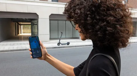 Femme regardant son téléphone, une Segway-Ninebot E2 Pro E garée dans la rue derrière elle