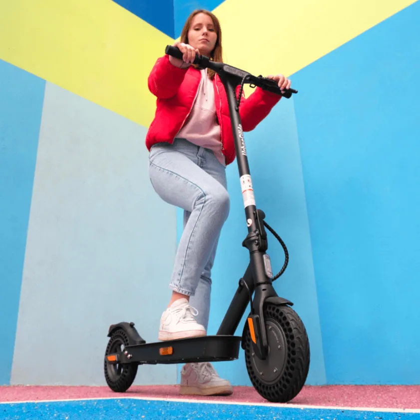 Une jeune femme en veste rouge debout sur l'Urbanglide Ride 100XS devant un mur bleu et jaune