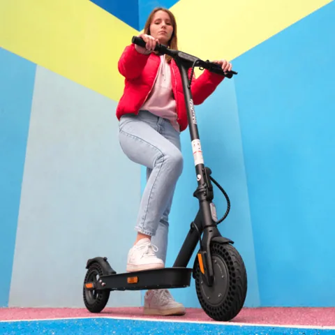 Une jeune femme en veste rouge debout sur l'Urbanglide Ride 100XS devant un mur bleu et jaune
