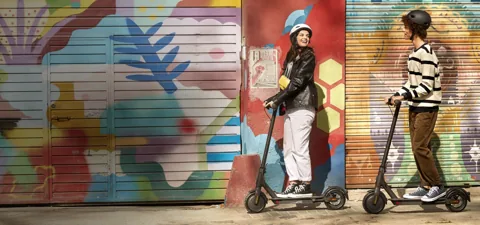 Deux personnes debout sur leur Mi Electric Scooter Essential noire devant un mur peint