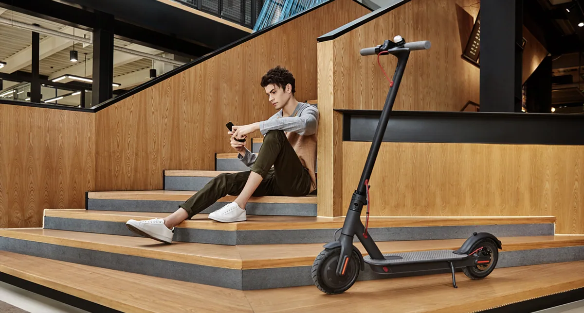 Mi Electric Scooter (M365) noire sur sa béquille près d'un homme assis sur des marches