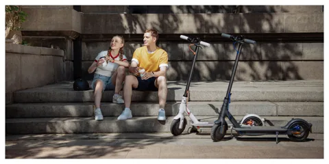 Deux Mi Electric Scooter 3 sur leur béquille près de deux personnes assises sur des marches